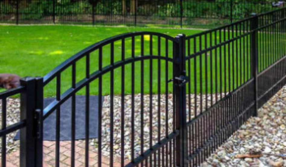 Black metal fence with an arched gate enclosing a yard with green grass, stone landscaping, and a brick pathway.