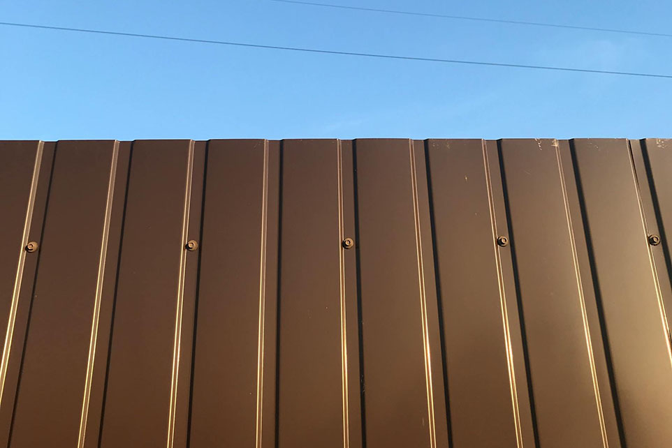 Metal brown fence panels with visible screws against a clear blue sky.