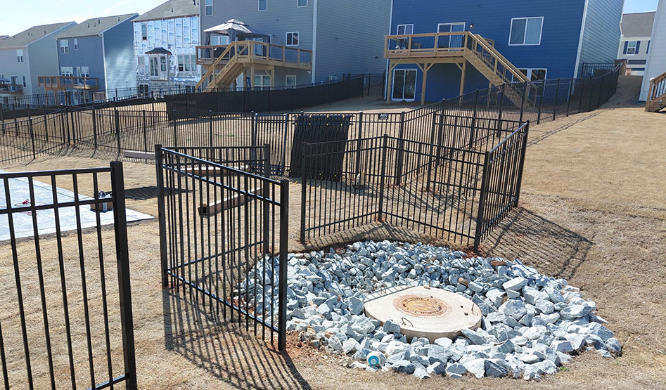 Black metal fence encloses a circular drainage area with rocks in a residential backyard with houses in the background.